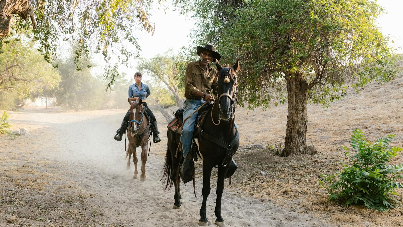 Two people riding horses on a rural dirt path surrounded by trees in a peaceful countryside setting.