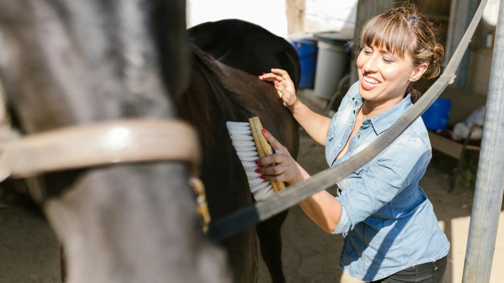 Smiling woman brushing a horse in a sunny outdoor stable, emphasizing care and connection.