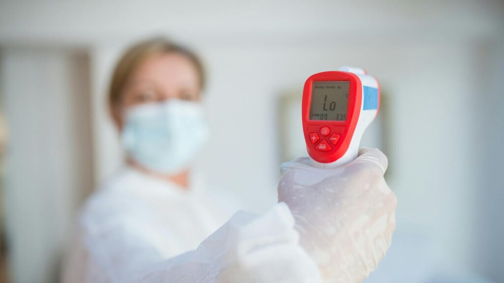 Masked healthcare worker using an infrared thermometer indoors for temperature screening.