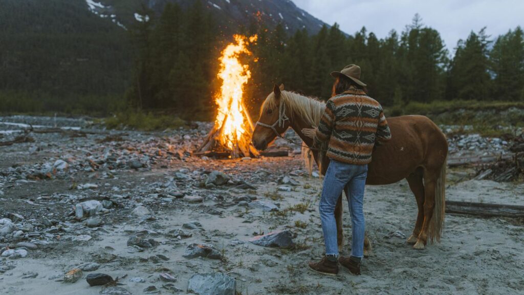 A man in a patterned sweater stands with a horse by a campfire in a scenic mountainous area.