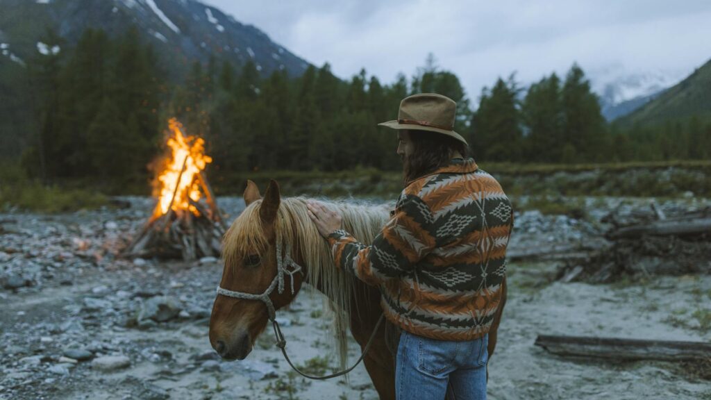 A man in a patterned jacket and hat pets a horse near a campfire set against a mountainous backdrop.