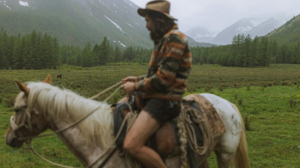 A man riding a horse through a scenic mountain valley with lush greenery and majestic mountains.