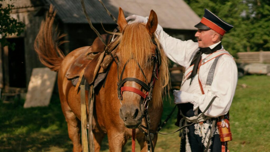 A man in a military uniform stands with a saddled horse outdoors.