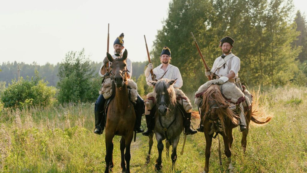 Three men in historical attire on horseback with rifles in a grassy field.