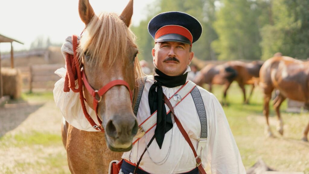 A vintage military horseman in period uniform stands beside a horse outdoors under a bright sky.