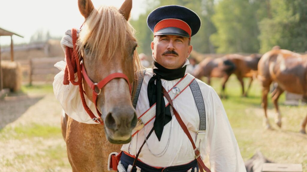A vintage military horseman in period uniform stands beside a horse outdoors under a bright sky.
