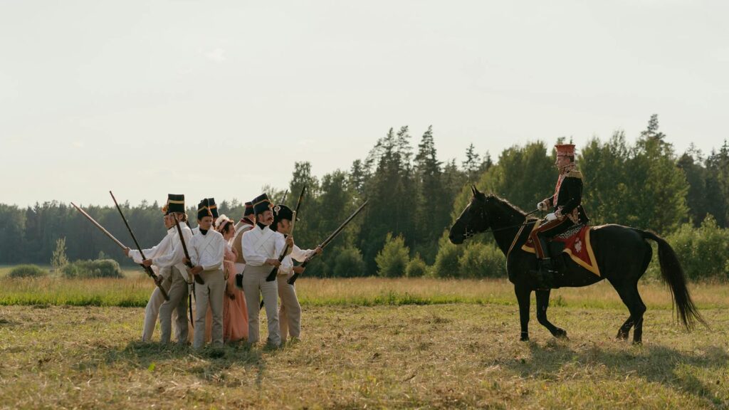 Men in historical uniforms and soldier on horseback reenact a battle scene outdoors.