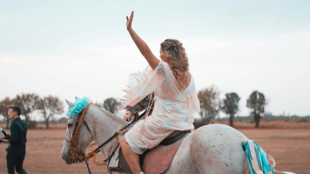 Woman riding a decorated white horse, waving amidst a scenic rural landscape during day.