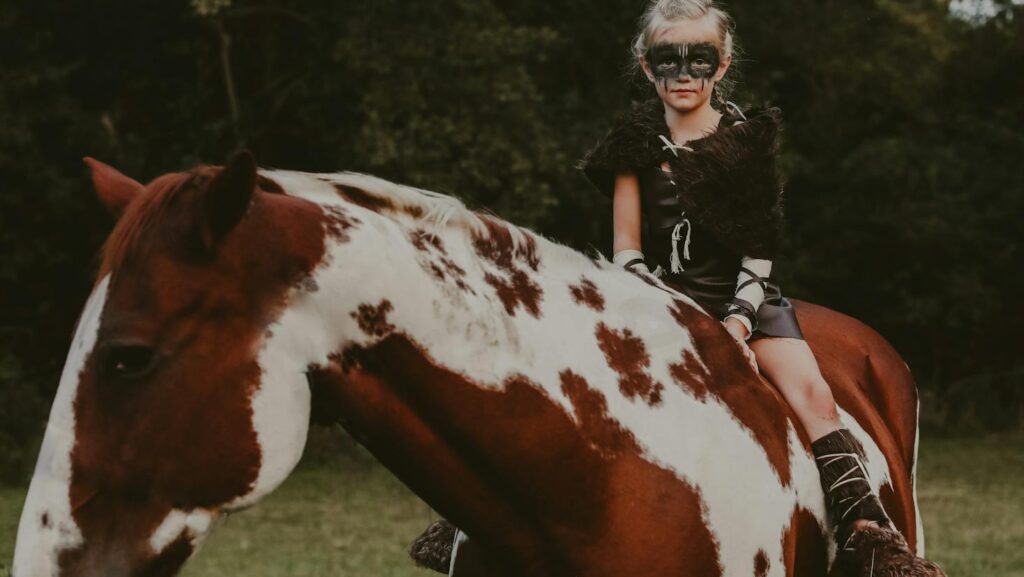 A child with face paint sits on a paint horse in a forested area, dressed in costume.