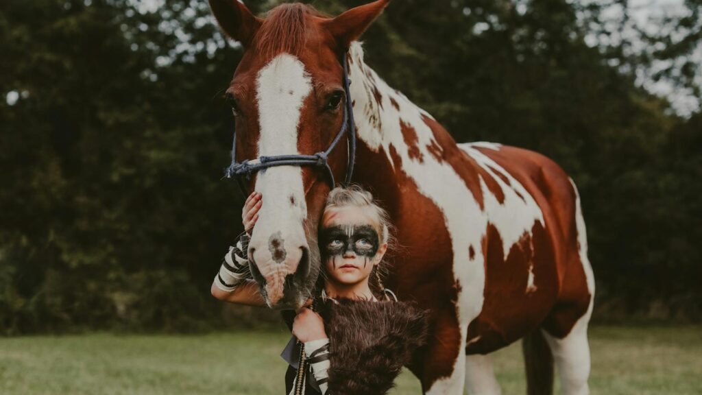 A child dressed as a Viking warrior with face paint stands beside a brown and white horse.