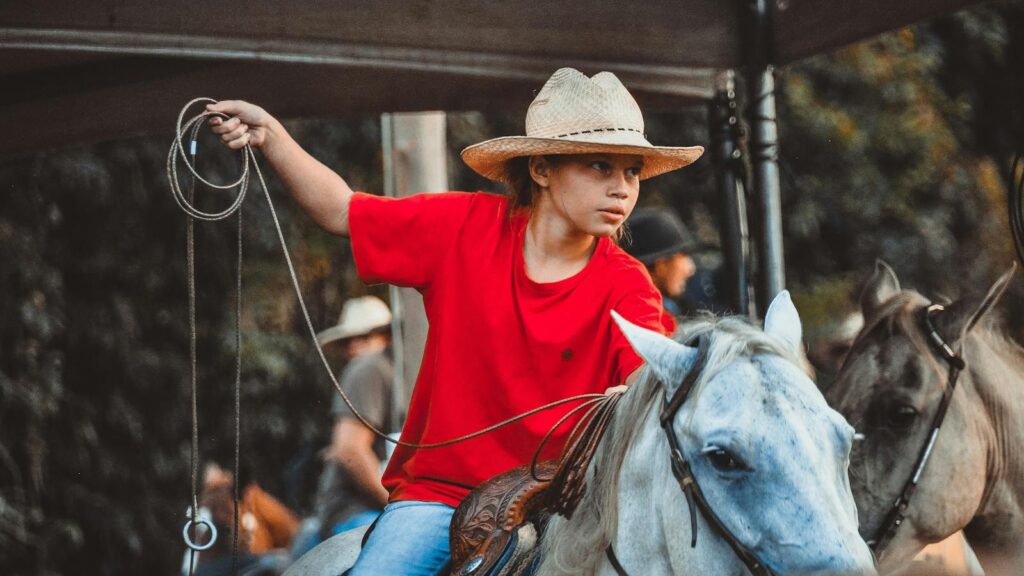 A child in a cowboy hat and red shirt skillfully rides a white horse, holding a lasso.