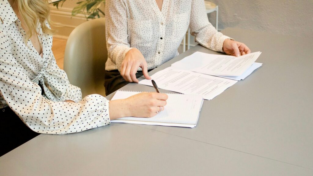 woman signing on white printer paper beside woman about to touch the documents