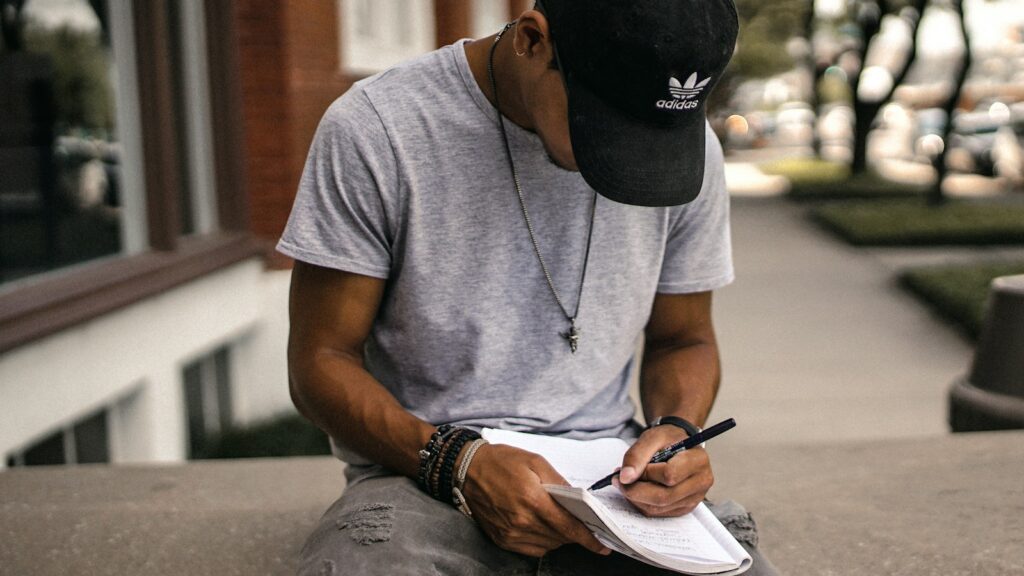 person in black adidas cap sitting on bench writing on notebook