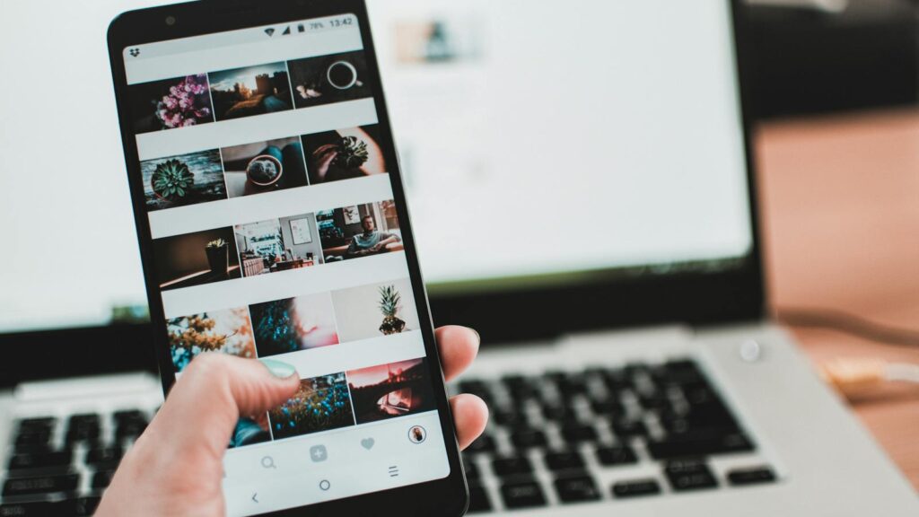 A person uses a smartphone to browse social media in front of a laptop, emphasizing modern technology and lifestyle.