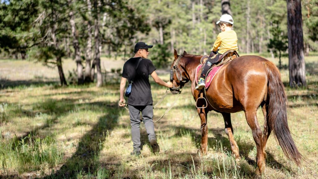 A child receives a horseback riding lesson in a scenic forest, led by an instructor during a sunny day.