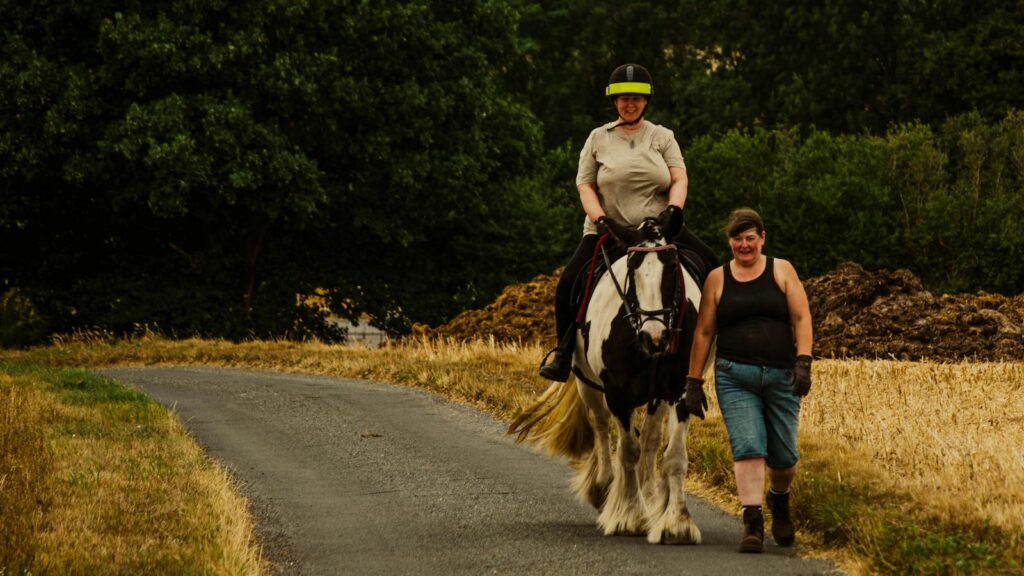 Two women enjoying a horse ride in the countryside on a quiet rural road, surrounded by nature.
