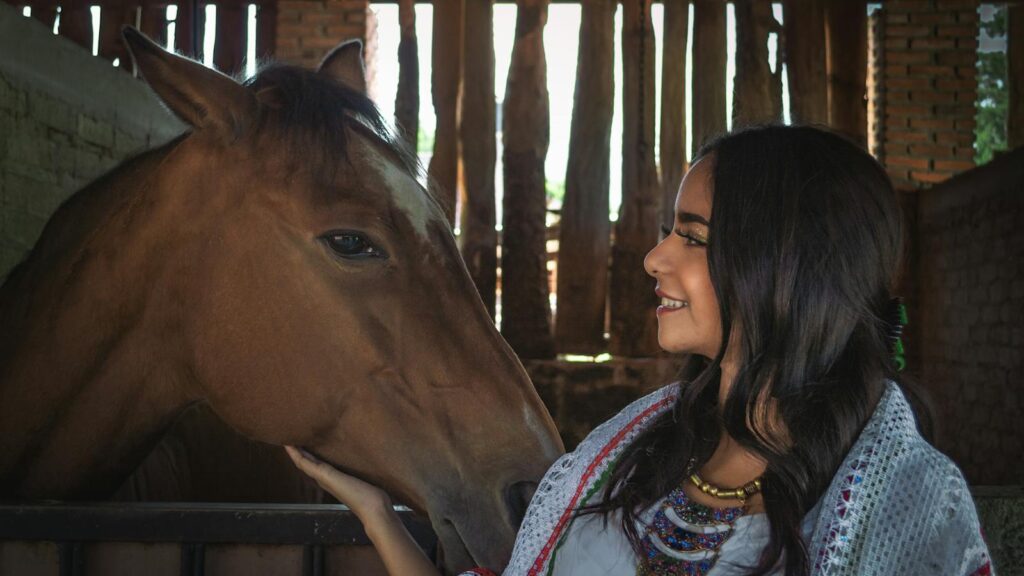 A woman smiles warmly while patting a horse inside a stable.