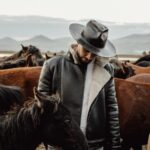 A cowboy in a leather jacket and hat stands among a herd of horses in a scenic outdoor field with mountains in the background.