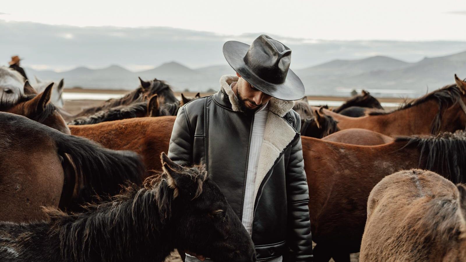 A cowboy in a leather jacket and hat stands among a herd of horses in a scenic outdoor field with mountains in the background.