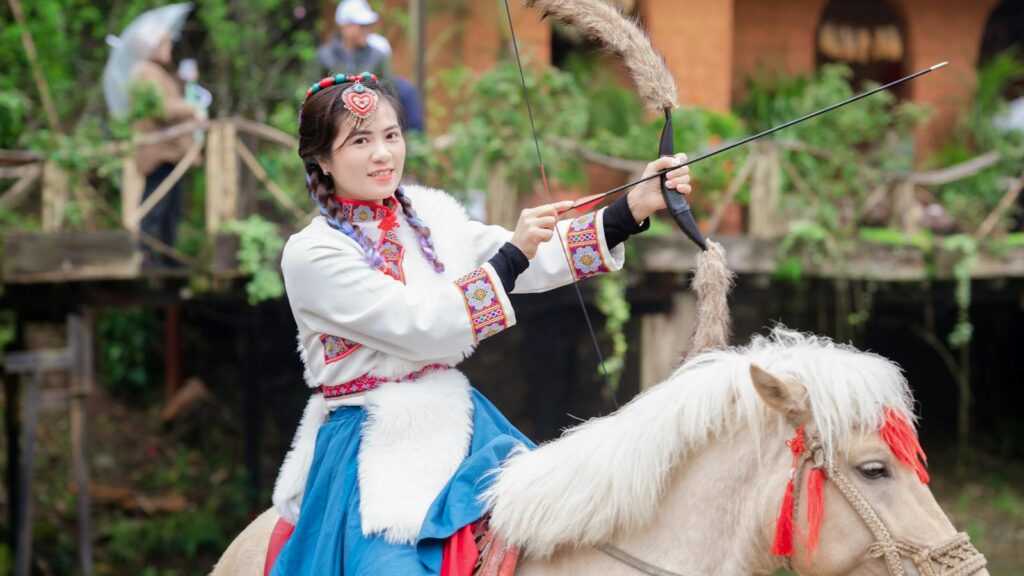 Woman wearing traditional attire on horseback, holding a bow outdoors.