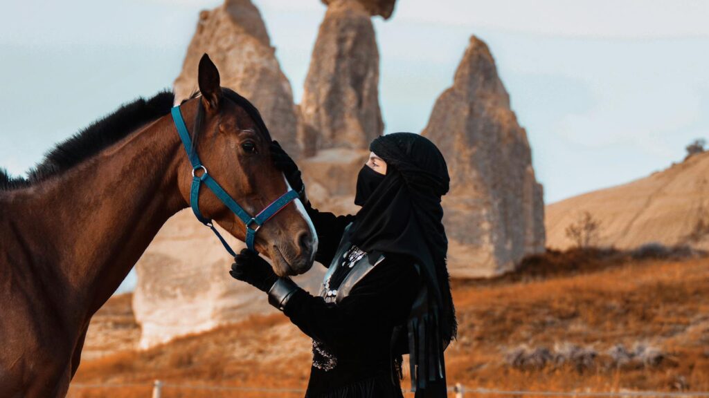 A woman in traditional attire with a horse against a rocky desert landscape.