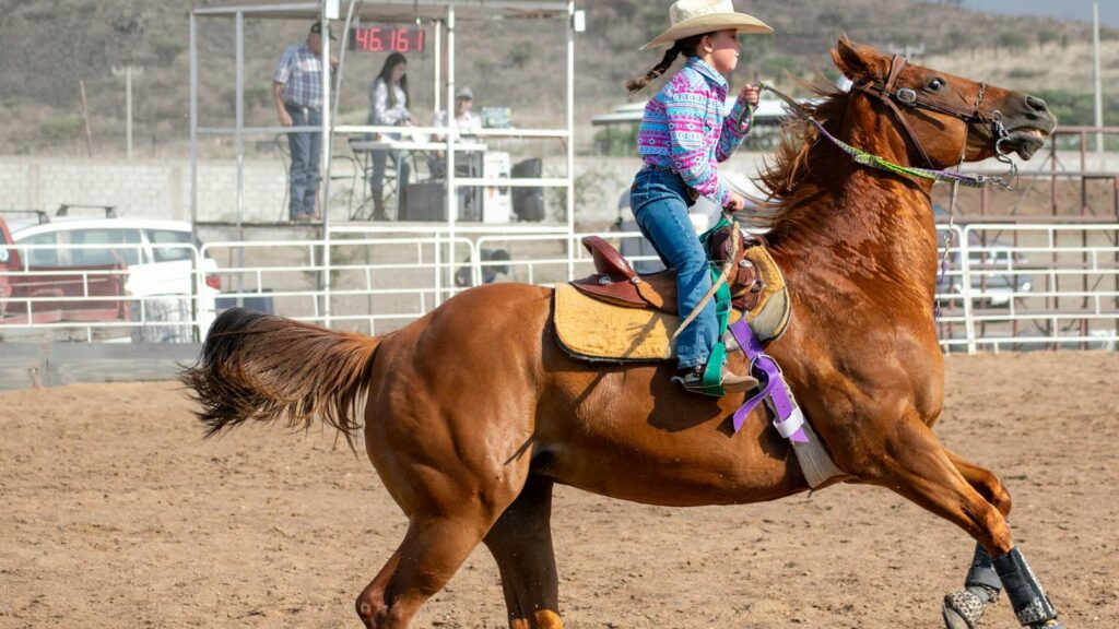 A young cowgirl rides a horse during a rodeo competition at an outdoor arena.