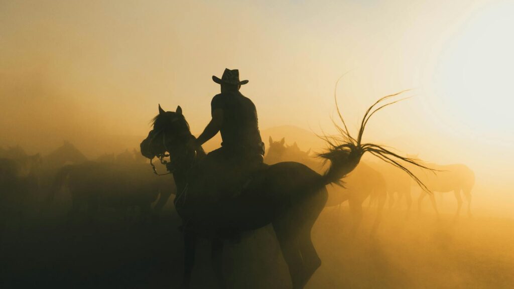 A cowboy riding a horse through a dusty sunrise, creating a dramatic silhouette.