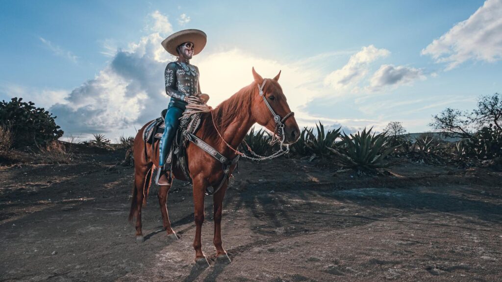 A man in traditional Mexican attire riding a horse in a rural landscape under a bright blue sky.