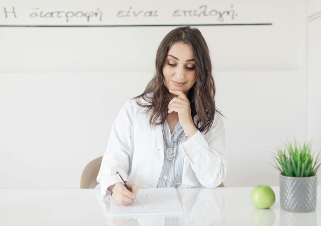A female nutritionist in a lab coat writing notes with an apple and plant on the desk.