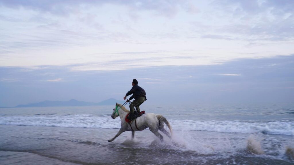 A man rides a galloping horse along a serene beach, with waves splashing under a cloudy sky.