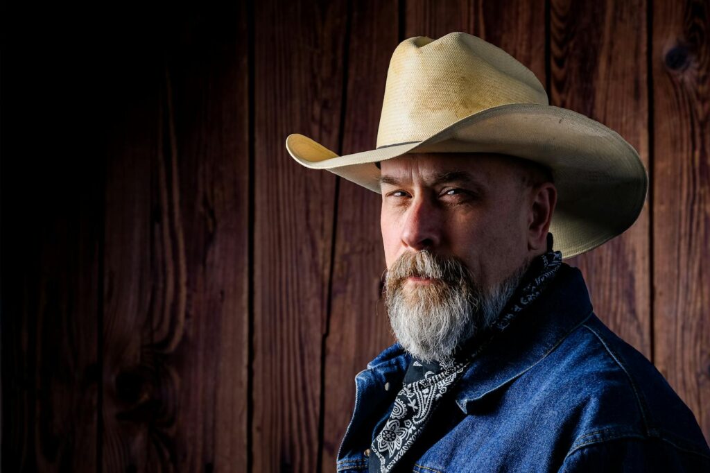 Rugged cowboy with a beard and hat posing against a rustic wooden background.