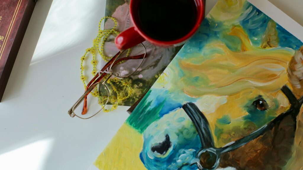 A vibrant still life featuring a coffee cup, colorful horse painting, and eyeglasses on a table.