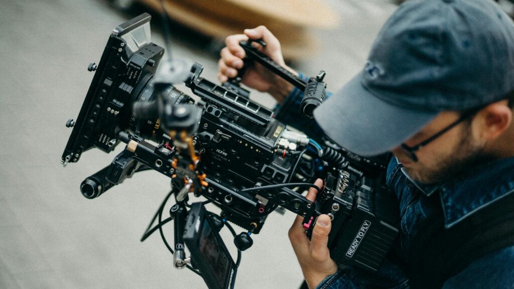 A man with a cap operates a professional video camera outdoors during daytime, capturing cinematic footage.