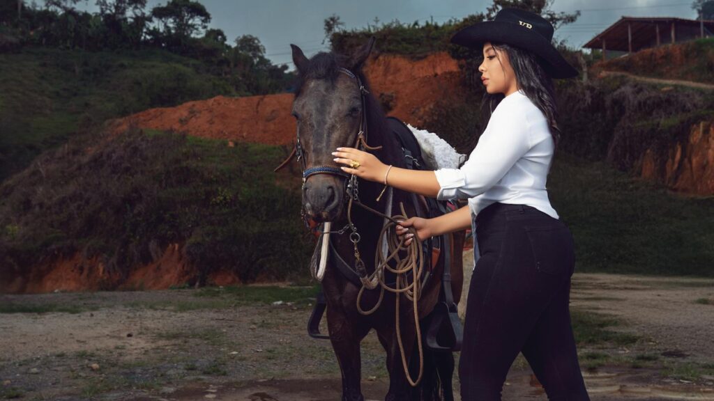 A young woman in Western attire standing beside a horse in a hilly outdoor setting.