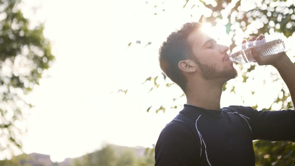 Side view of a young man drinking water from a plastic bottle outdoors in sunlight.