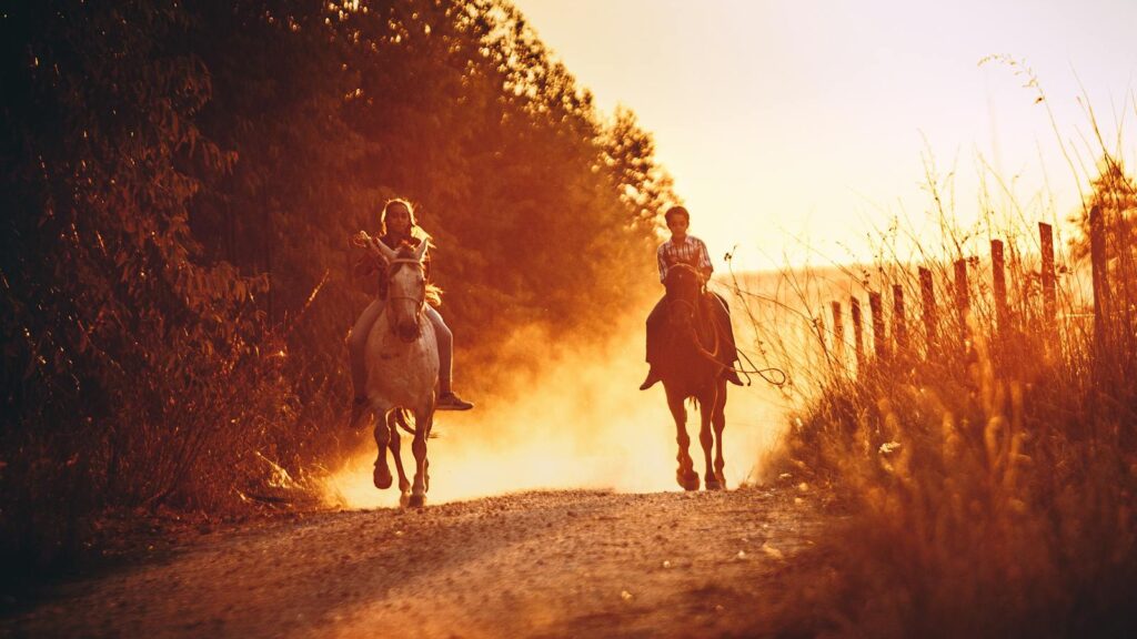 Two children horseback riding on a dusty road at sunset, creating a warm, serene atmosphere.