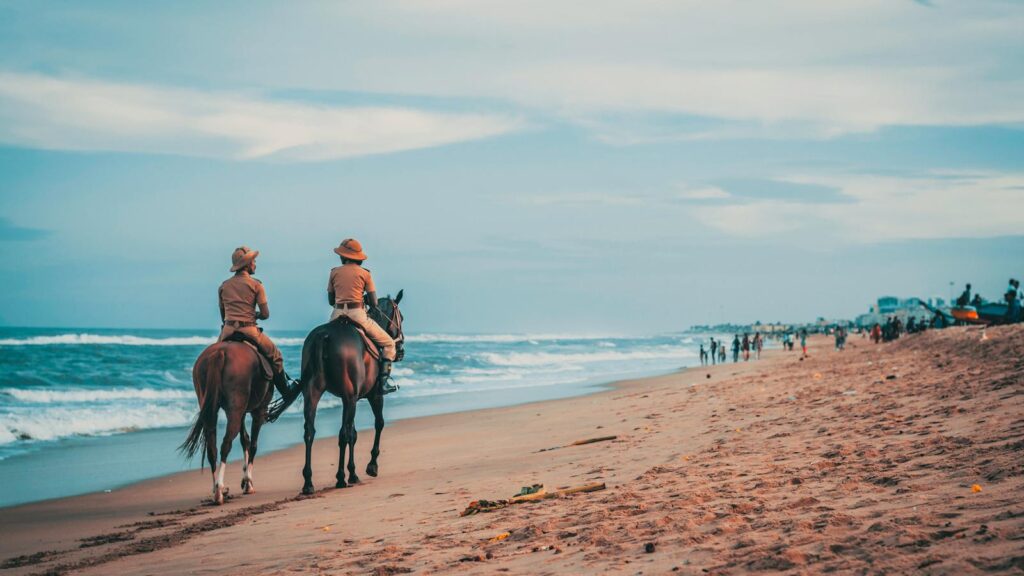Two riders on horseback enjoying a peaceful ride along a scenic beach with waves and cloudy sky.