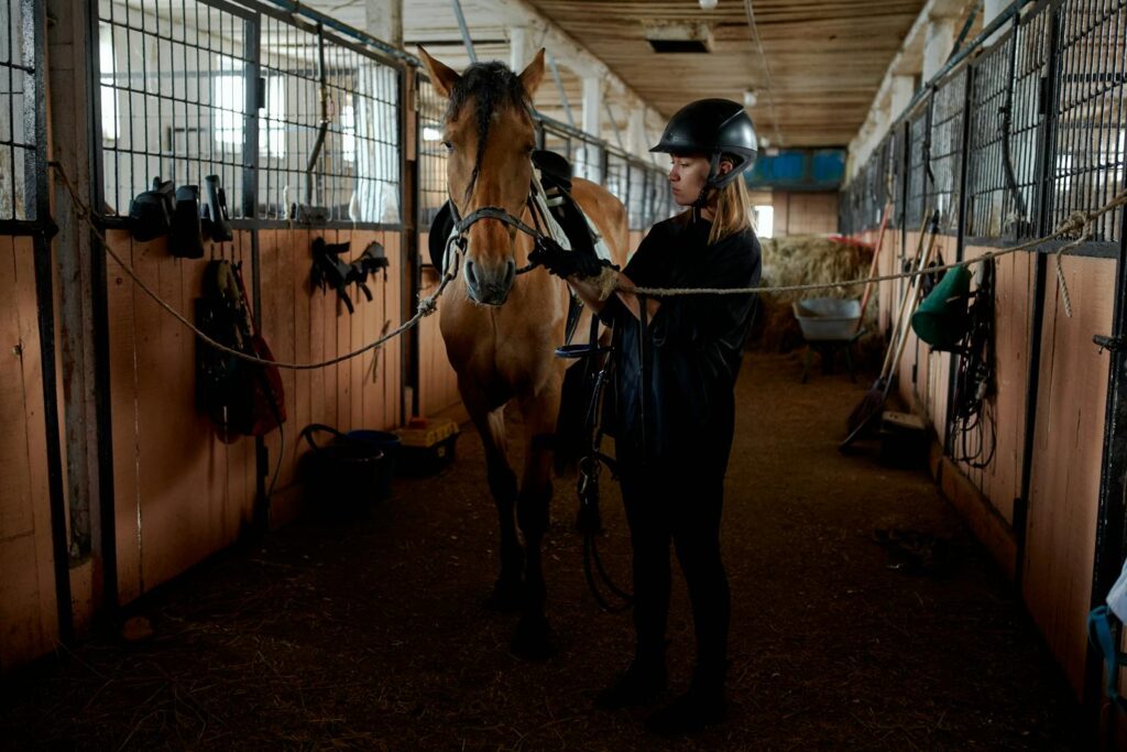 A woman in riding gear prepares a horse in an indoor stable setting, focusing on equestrian care.