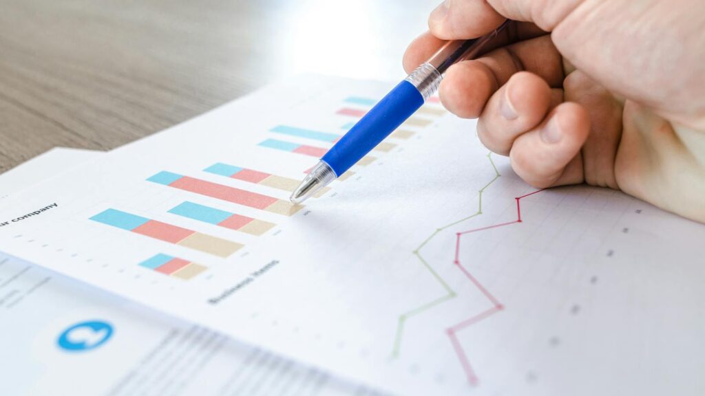 A close-up of a hand with a pen analyzing data on colorful bar and line charts on paper.