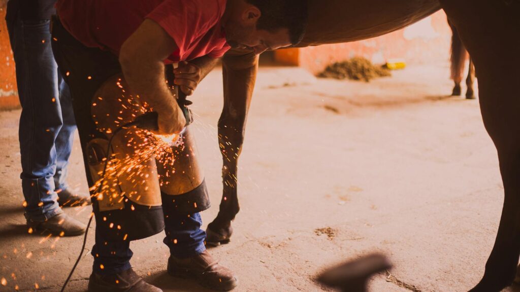 A farrier shoeing a horse in an indoor stable with flying sparks from the equipment.