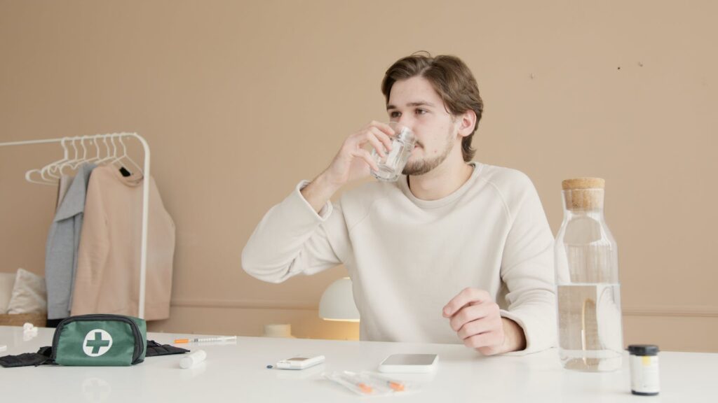 Man in white long sleeves drinking water from glass at home with medical supplies.