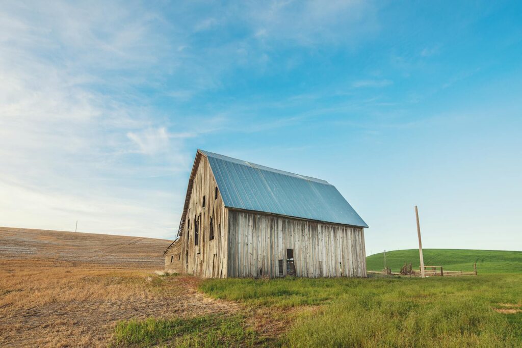 A rustic wooden barn stands in a serene rural countryside under a clear blue sky.
