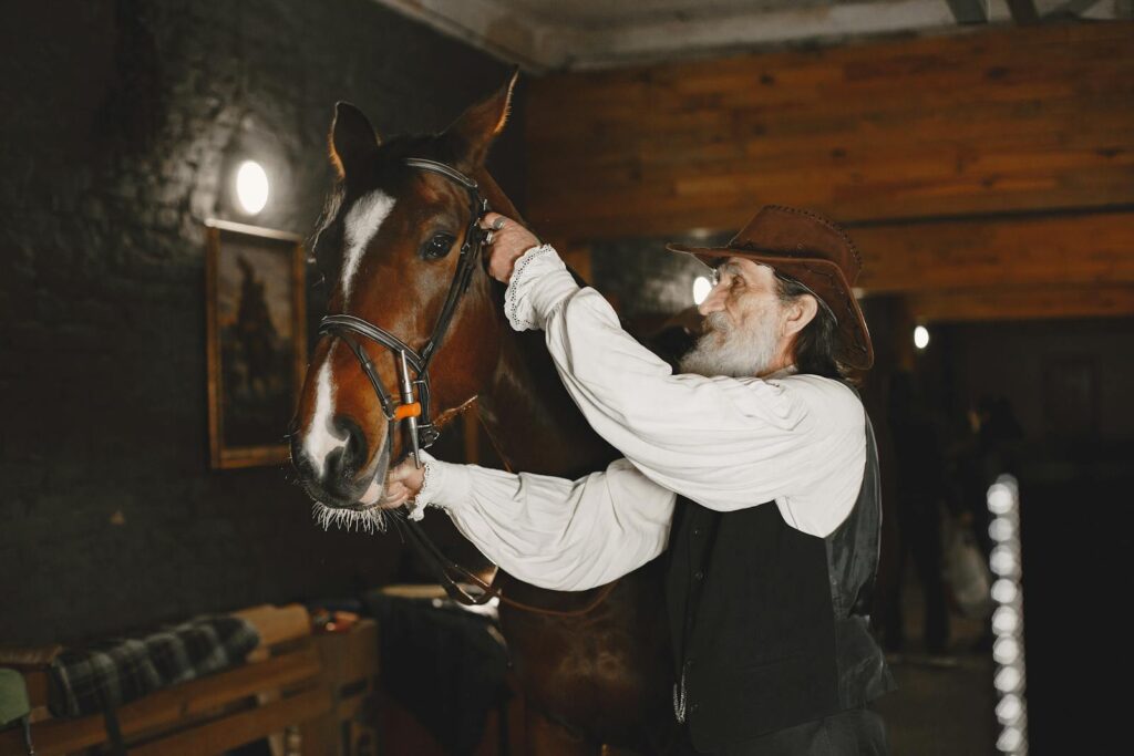 An elderly cowboy gently tends to a brown horse inside a stable, emphasizing human-animal connection.