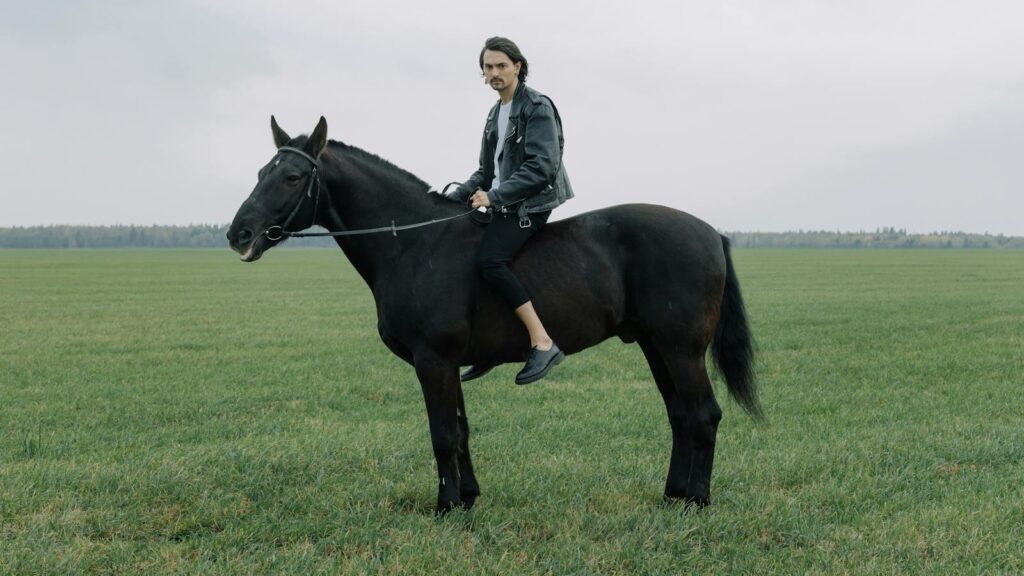 A man riding a black horse in a vast green field on an overcast day.