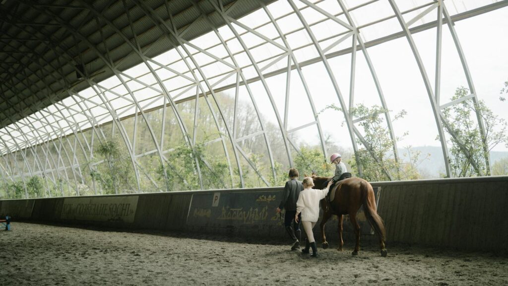 Child learning horseback riding in an indoor equestrian arena with guidance from instructors.