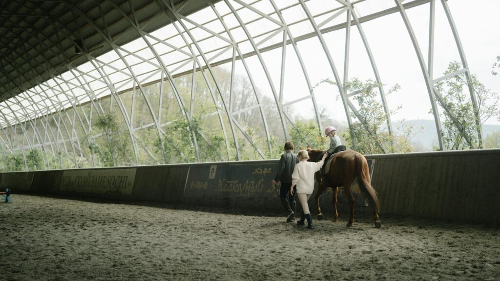 Child learning horseback riding in an indoor equestrian arena with guidance from instructors.