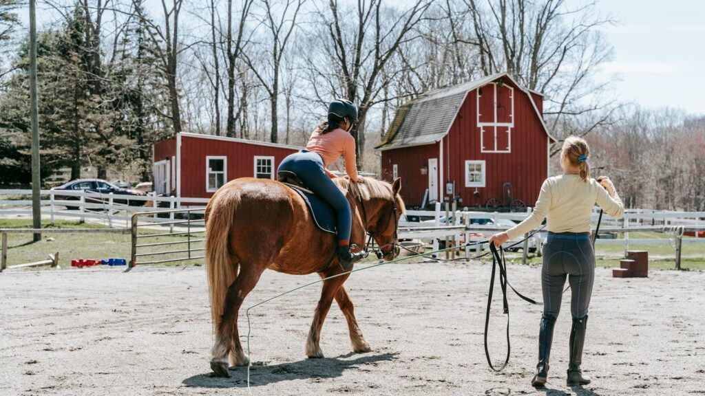 A woman trains a horse rider outdoors near a red barn on a sunny day.