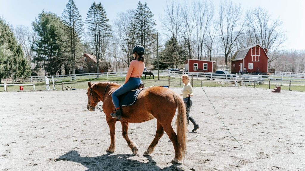 A woman learning horseback riding in an outdoor arena with instructor guidance.