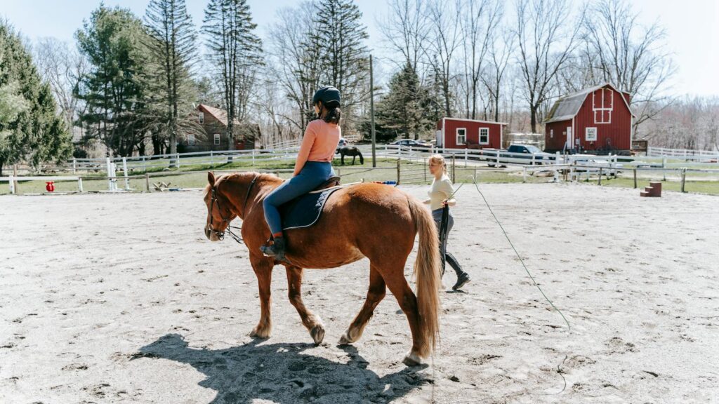 A woman learning horseback riding in an outdoor arena with instructor guidance.