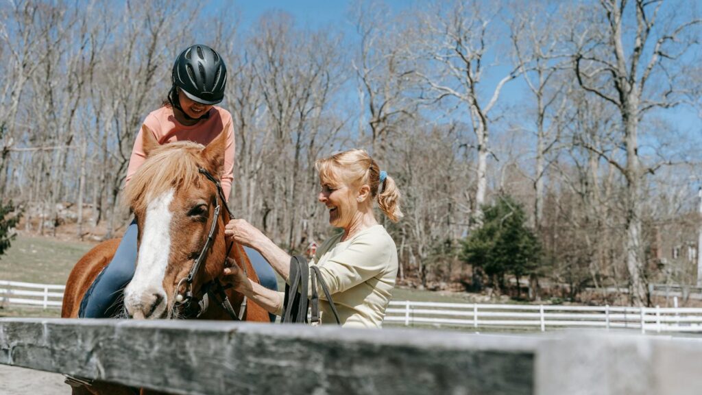 A woman giving a horse riding lesson to a child in a sunny outdoor arena, emphasizing safety and fun.
